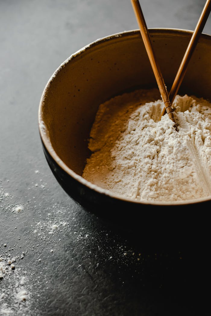 A close-up of flour in a ceramic bowl with chopsticks on a dark surface.